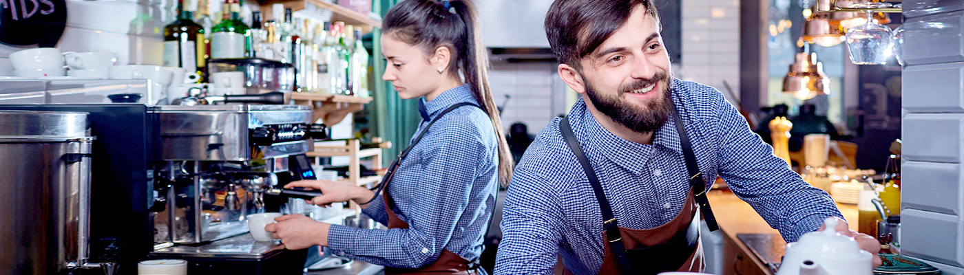 Zwei Baristas in einem Café, einer bedient eine Kaffeemaschine, während der andere eine Tasse hält.